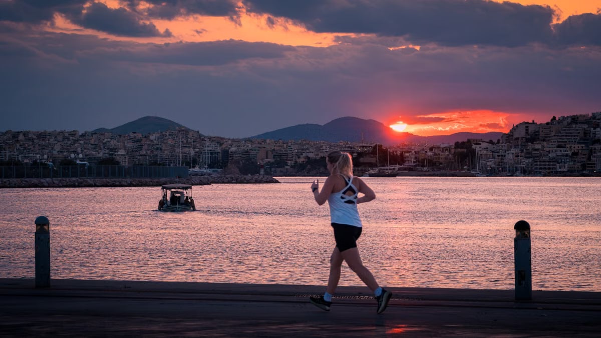 A woman jogs along a waterfront at sunset with a boat in the background.